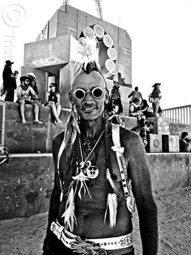 burning man - man in costume near man base, attire, burning man outfit, feathers, headdress, headwear, mahawk, native american, necklaces, neckless, sunglasses